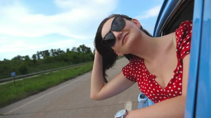 戴着太阳镜的年轻女孩斜靠在老式车窗外，享受旅行。女人从移动复古汽车中望出去。旅游概念。穿着旧车的迷人