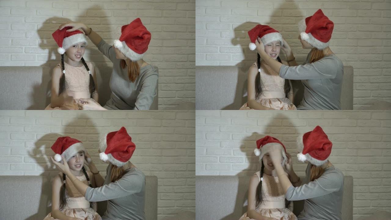 姐姐帮助小女孩将圣诞老人的帽子戴在头上。他们坐在家里的沙发上，靠着白色的砖墙微笑。
