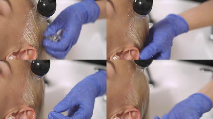 蓝色橡胶手套中的一只手的特写镜头，在反冲洗盆中清洗金发女郎的染发。洗完头发后冲洗干净