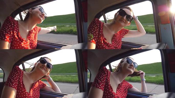 戴着太阳镜的快乐女孩斜靠在复古车窗外，享受旅行。年轻女子在阳光明媚的日子里看着窗外移动老式汽车。旅行