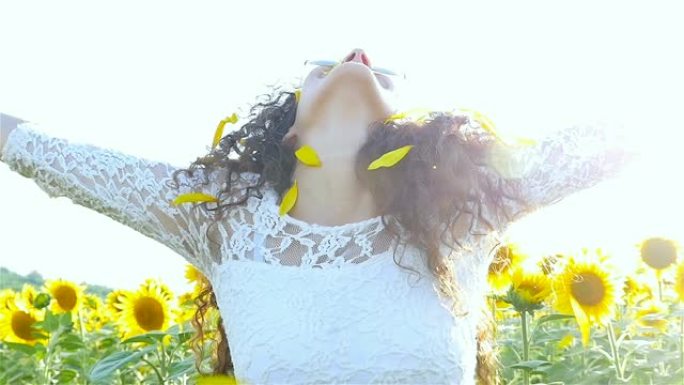 美丽的幸福女孩女人在日落时在向日葵田上头顶上撒播向日葵花瓣。自由概念。慢动作