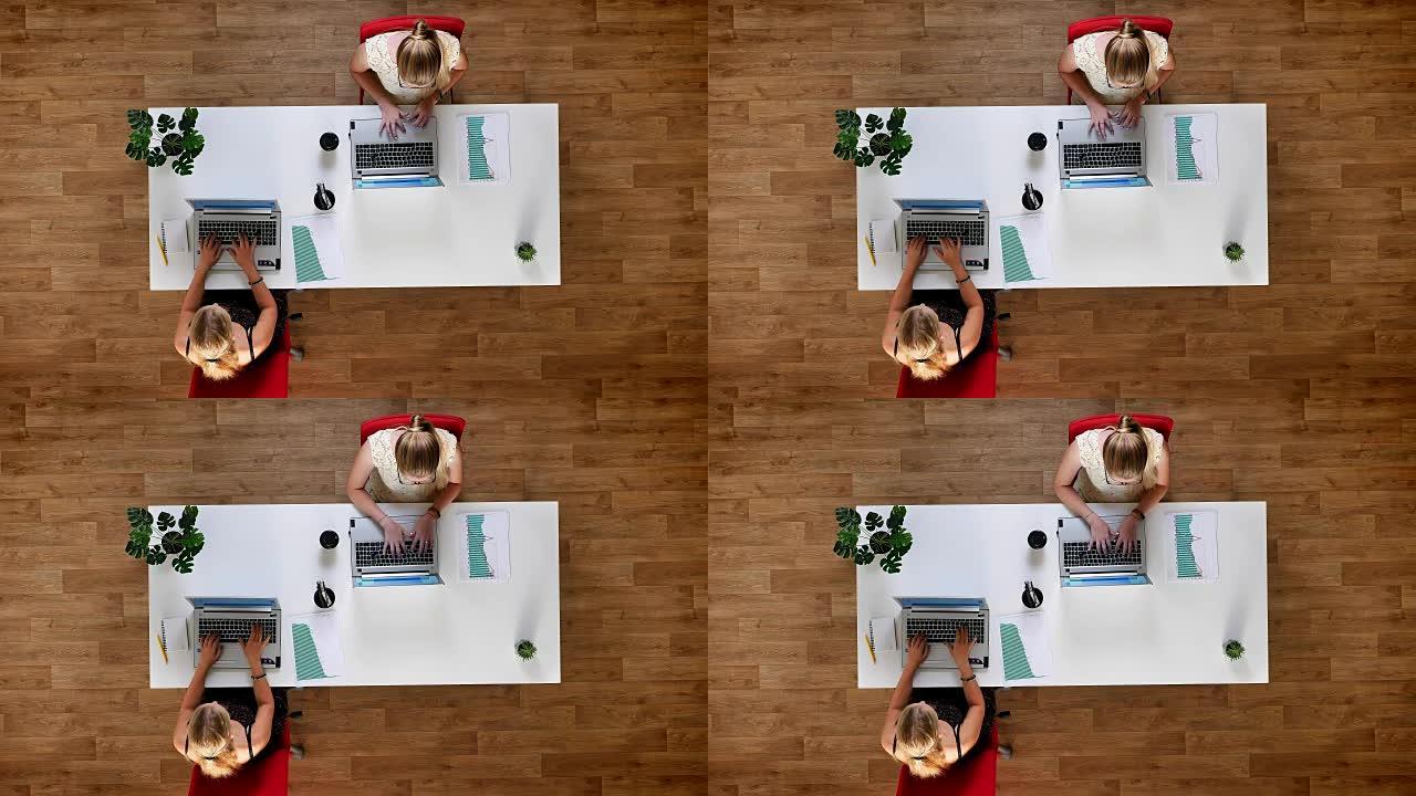 自上而下拍摄了富有成效的同事，两个女孩坐在公共桌子旁，在室内木地板上的笔记本电脑上工作