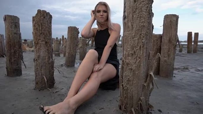 年轻的金发女子坐在盐峡湾海岸，周围是木桩，多云的天气
