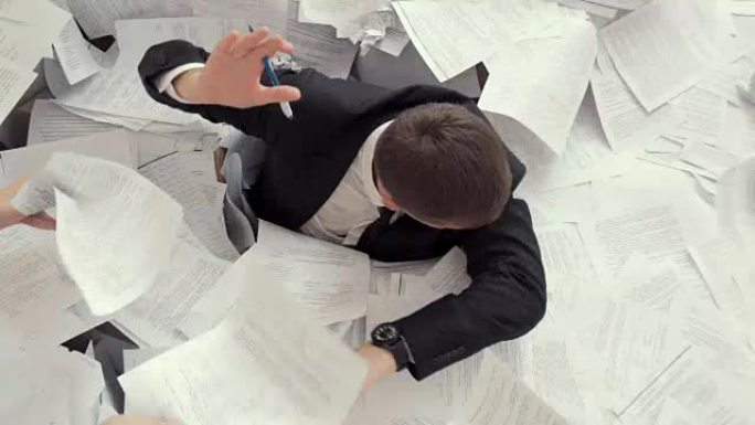 一个年轻的疲倦的男人坐在一堆文件中，签署了许多文件。办公室员工无法应付工作，看着时钟