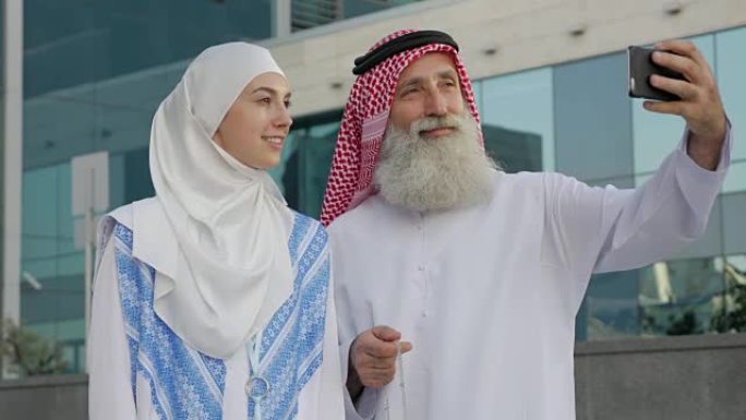 一个留着胡子的阿拉伯老年男子和一个年轻女孩在城市里用智能手机自拍