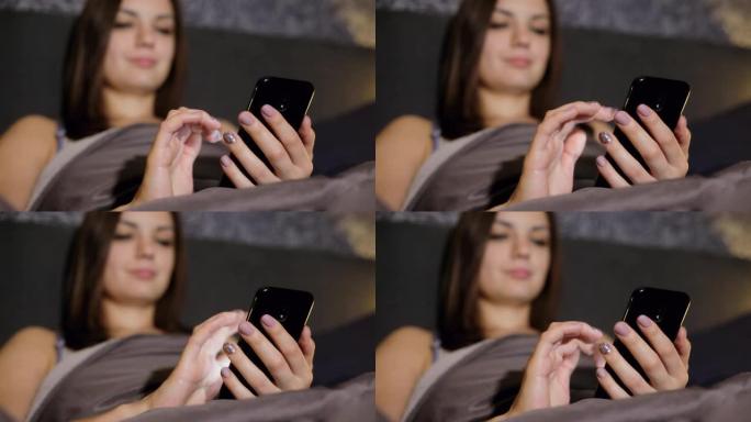 晚上躺在卧室的床上上网的女孩的特写肖像，写短信，看照片，聊天。专注于手机