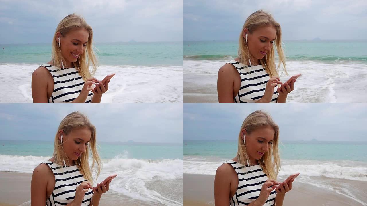 年轻漂亮的苗条女人，长长的金发，穿着黑白连衣裙，站在海岸上，在海上风暴中使用智能手机。海滩上的女孩触