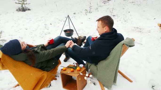 这对夫妇坐在舒适的沙发上的冬季森林的篝火旁。下雪。冬季快乐概念。