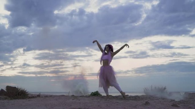 美丽的女孩，穿着粉红色的衣服，在河岸上的烟雾弹的雾中跳舞。一个有着花发型的性感女人的舞蹈。慢动作。