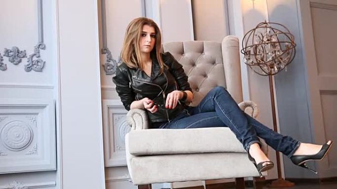 年轻女子穿着黑色皮夹克、牛仔裤和高跟鞋在时尚的扶手椅上摆姿势