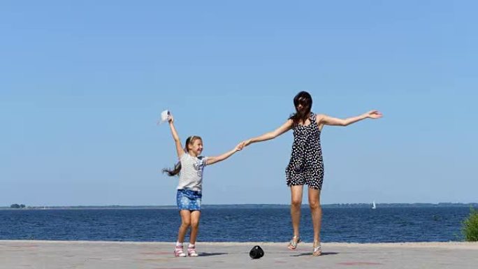 在海滩上，妈妈和女儿，穿着太阳裙的美丽黑发女郎和女童少年，在炎热的夏日跳舞，用迷你音乐蓝牙便携式扬声