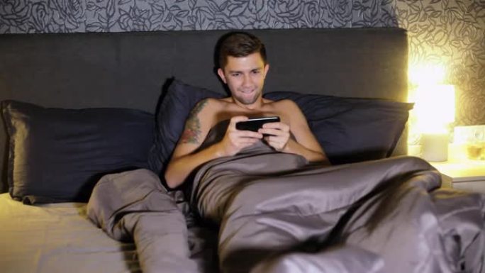年轻人晚上躺在家里的床上在智能手机上玩游戏。紧张地比赛，赢得比赛，非常高兴