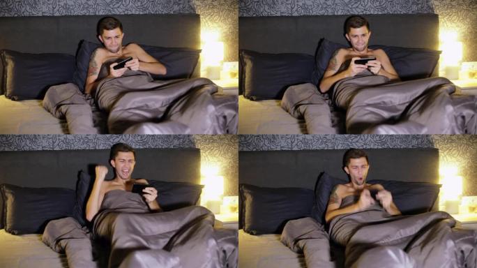 年轻人晚上躺在家里的床上在智能手机上玩游戏。紧张地比赛，赢得比赛，非常高兴