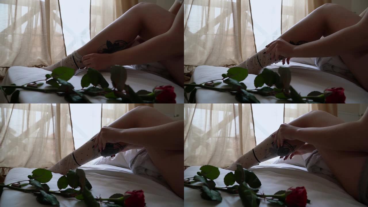 女人在带有纹身的腿上放了白色斑点的长袜。红玫瑰躺在女孩附近的床上。