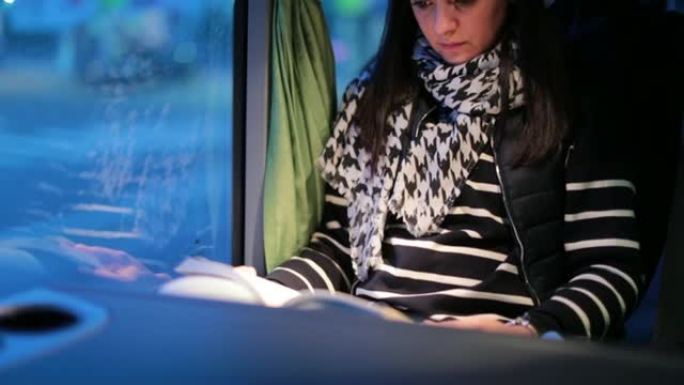 晚上乘公共汽车旅行时低头看书的女人。路过窗户的风景