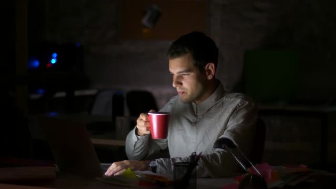 英俊的白人男性坐在桌面上，在笔记本电脑上工作，在黑暗的办公室室内时，屏幕上闪闪发光，从杯子里喝水的镜