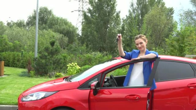 快乐的年轻女子是红色汽车的主人。她站着，靠在门上。年轻女子用钥匙挥手。她看起来很开心。