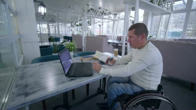 在线教育，成功的学生轮椅上的无效男子使用现代笔记本电脑技术学习在线教学和书籍在笔记本上做笔记特写坐在