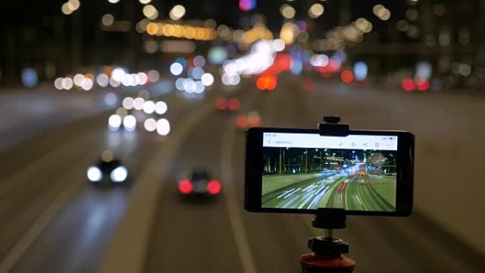 智能手机安装在三脚架上。他们在美丽的灯光背景下拍摄汽车运动的照片。晚上。