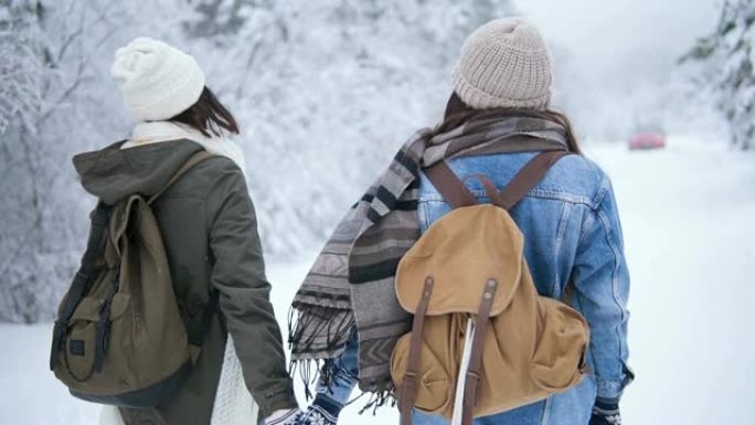 冬天，两个微笑的女人在公园散步。背着背包的朋友穿过森林。在白雪皑皑的森林背景下，穿着针织帽和冬装的女