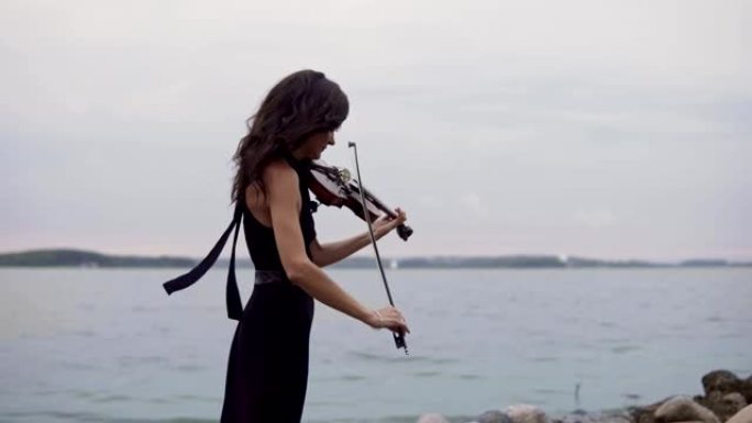 穿着黑色连衣裙的漂亮女孩在海上背景拉小提琴。4k中的艺术概念