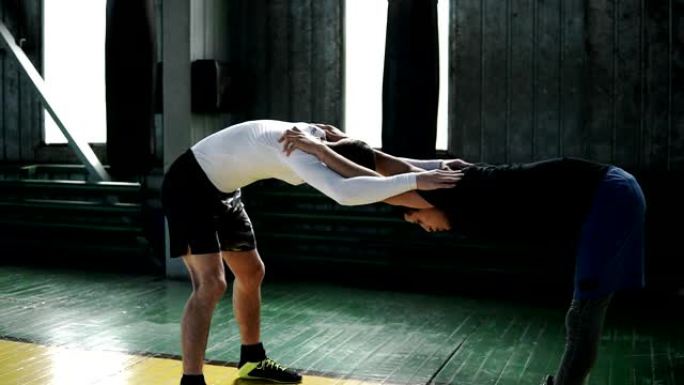 两名从事拳击运动的男子在健身房里通过互相倾斜和支撑来伸展手臂和背部，伸出双手放在肩膀上。前视图