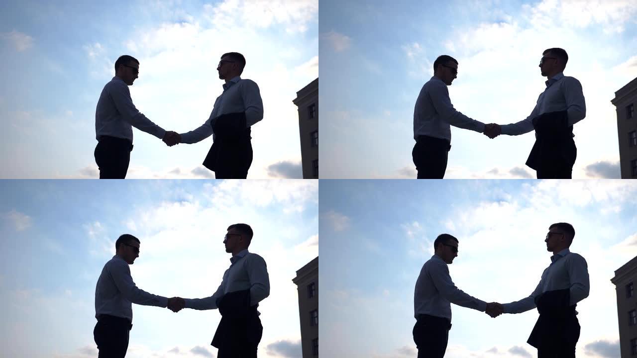 两个英俊的商人在城市环境中互相打招呼。年轻的男同事在城市握手，背景是蓝天。户外商业伙伴握手。慢动作