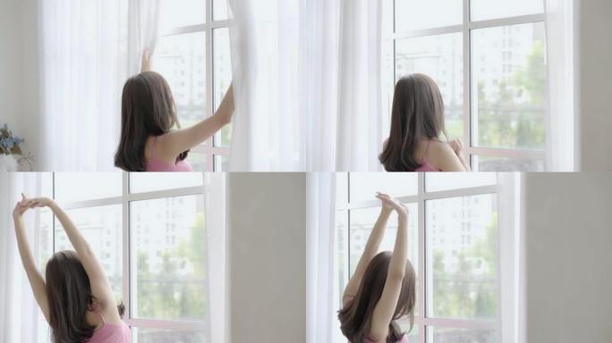 美丽的年轻亚洲女人醒来，早上带着打开的窗帘走到窗前，女孩在假期中带着幸福，生活方式理念，慢动作放松。