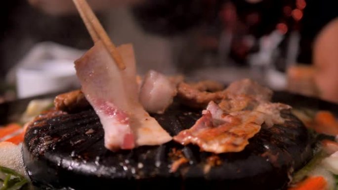 人们用筷子做饭，拿起和翻转猪肉，并在深色和谷物加工的火锅上遇到韩国烧烤烧烤