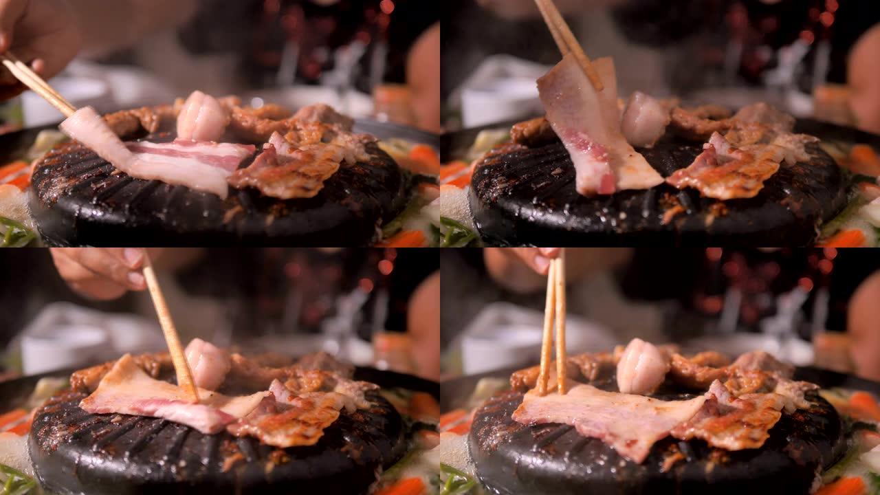 人们用筷子做饭，拿起和翻转猪肉，并在深色和谷物加工的火锅上遇到韩国烧烤烧烤