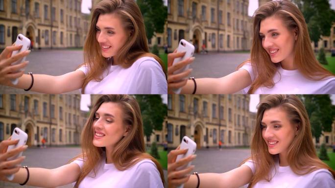 阳光灿烂的日子，害羞而漂亮的长发白人女孩在城市景观上用她的设备自拍
