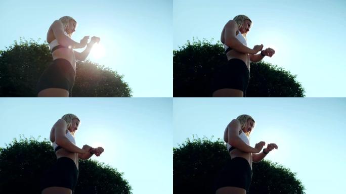 低角度运动的年轻金发女子完成慢跑，站在阳光下，背靠天空背景，检查她的智能手表的心率和速度