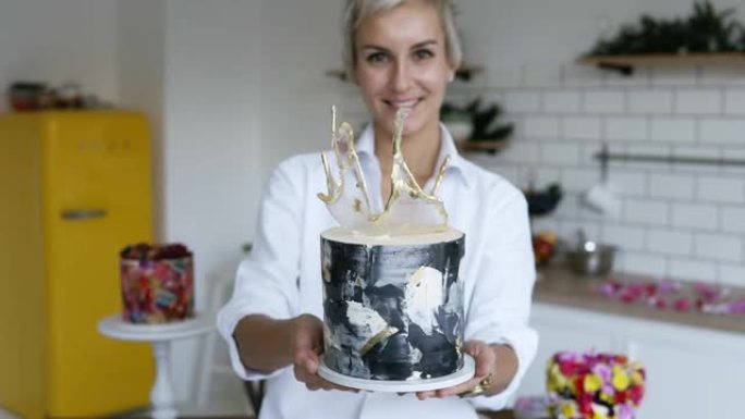 穿着白衬衫的女性糖果店在她的厨房里展示了现代装饰的黑白蛋糕。黄色冰箱和白色墙壁厨房，背景上装饰精美的