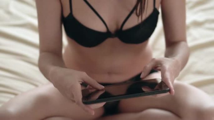 穿着黑色内衣的年轻女子坐在床上兴致勃勃地玩着平板电脑