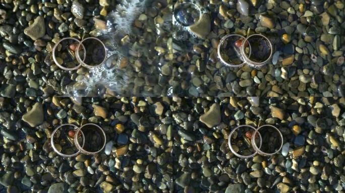 两枚金色结婚戒指躺在沙滩上的石头上，照耀着阳光，特写宏观。戒指上的光输血。海浪冲在他们身上