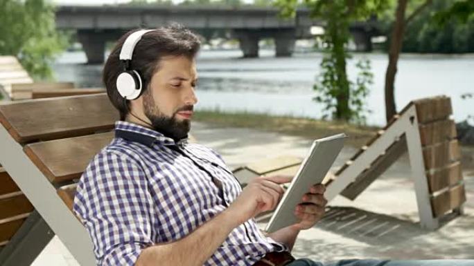 盖伊躺在长凳上，通过耳机听音乐。他还拿着平板电脑在屏幕上打字。男人正在看屏幕并移动他的发放者。他上下