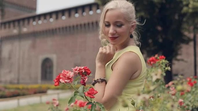 穿着黄色连衣裙的美丽金发女郎站在米兰盛开的玫瑰灌木丛旁边