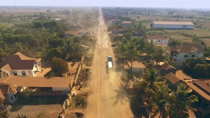 空中无人机射击: 在尘土飞扬的道路上飞越穿越小村庄的车辆