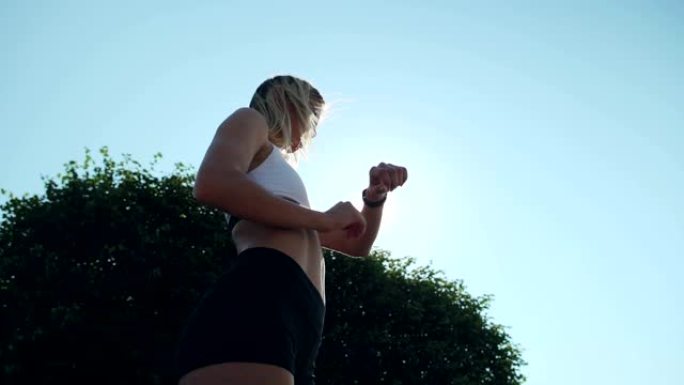 穿着运动胸罩和短裤的自信女运动员的低角度站在阳光下的绿树旁，在跑步训练前设置智能手表，她的金发在风中