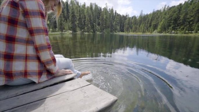 金发女孩坐在湖畔，在水中享受大自然。女人在高山湖泊的水中泼水。慢动作