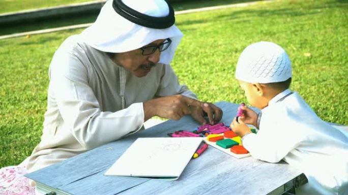 手持视图: 穆斯林祖父教儿子早上在阳光下在公园绘画