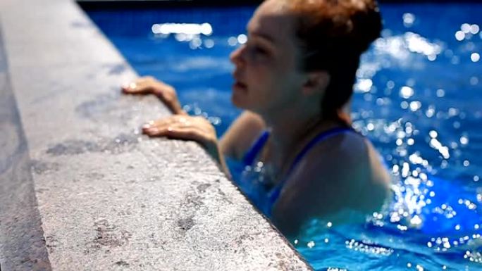 一个年轻漂亮的女人，红头发，蓝色泳衣，在阳光明媚的夏日在豪华的室外游泳池游泳。