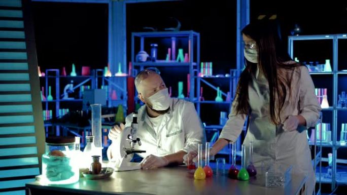 男性化学家在显微镜下观察反应，将移液器溶液吸到一块玻璃上。助手从烧瓶中倒出液体。