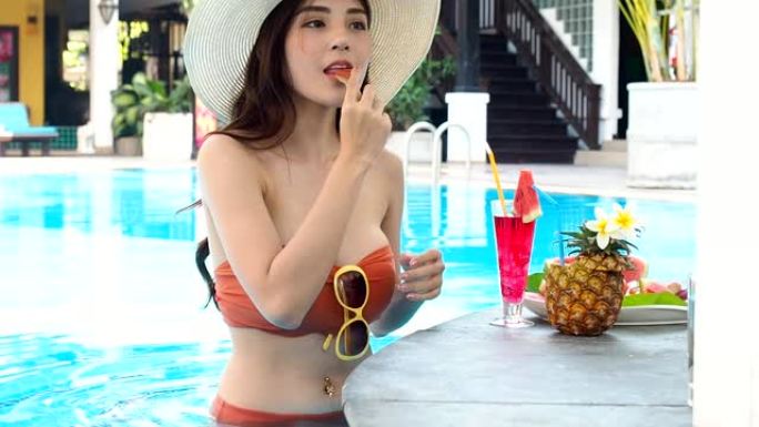 夏季概念。穿着橙色比基尼的性感亚洲女性在游泳池放松时吃玫瑰苹果。