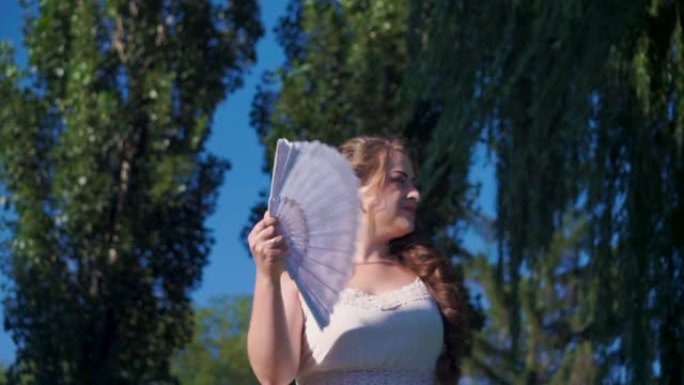 年轻女子挥舞着白色风扇看着太阳在炎热的夏日扇风
