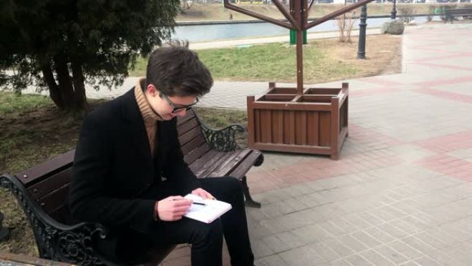 一个年轻人坐在长凳上，在笔记本上素描。春天，天气凉爽，那家伙穿着一件黑色外套。