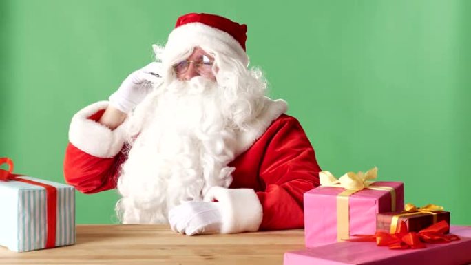 满意的圣诞老人在电话里说话，笑着，坐在桌子旁，拿着礼物，背景是绿色的chromakey。
