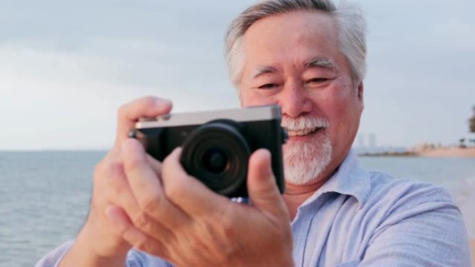 亚洲资深男子在海滩上使用相机，情绪愉快。有生活方式、退休和科技观念的人。