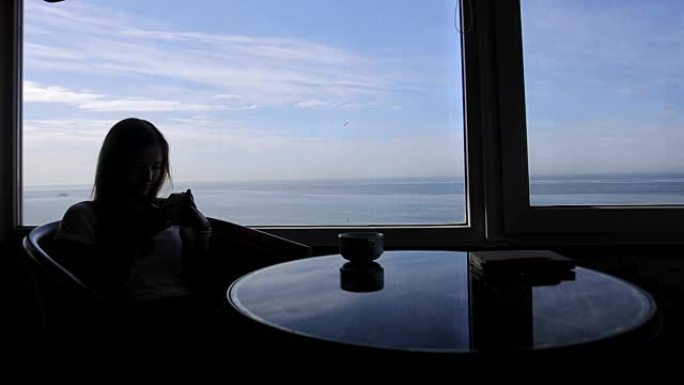 年轻女孩的轮廓的长镜头坐在咖啡馆里，可以看到大西洋的全景。早上用一杯咖啡，茶，书，记事本，使用智能手