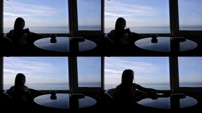 年轻女孩的轮廓的长镜头坐在咖啡馆里，可以看到大西洋的全景。早上用一杯咖啡，茶，书，记事本，使用智能手